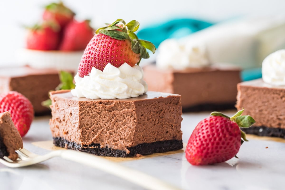 chocolate cheesecake square topped with whipped cream and a strawberry with one bite missing